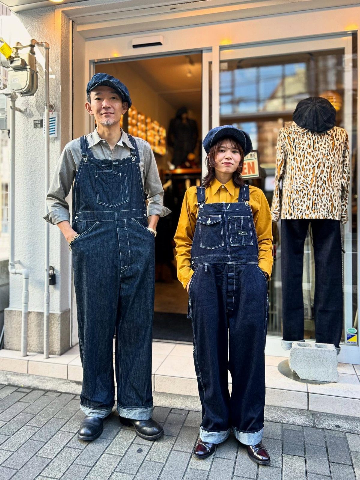 Man and woman wearing dark denim overalls and blue berets outside a store.