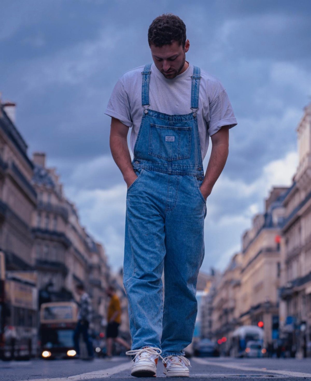 Man wearing blue denim overalls and a white t-shirt on a city street.