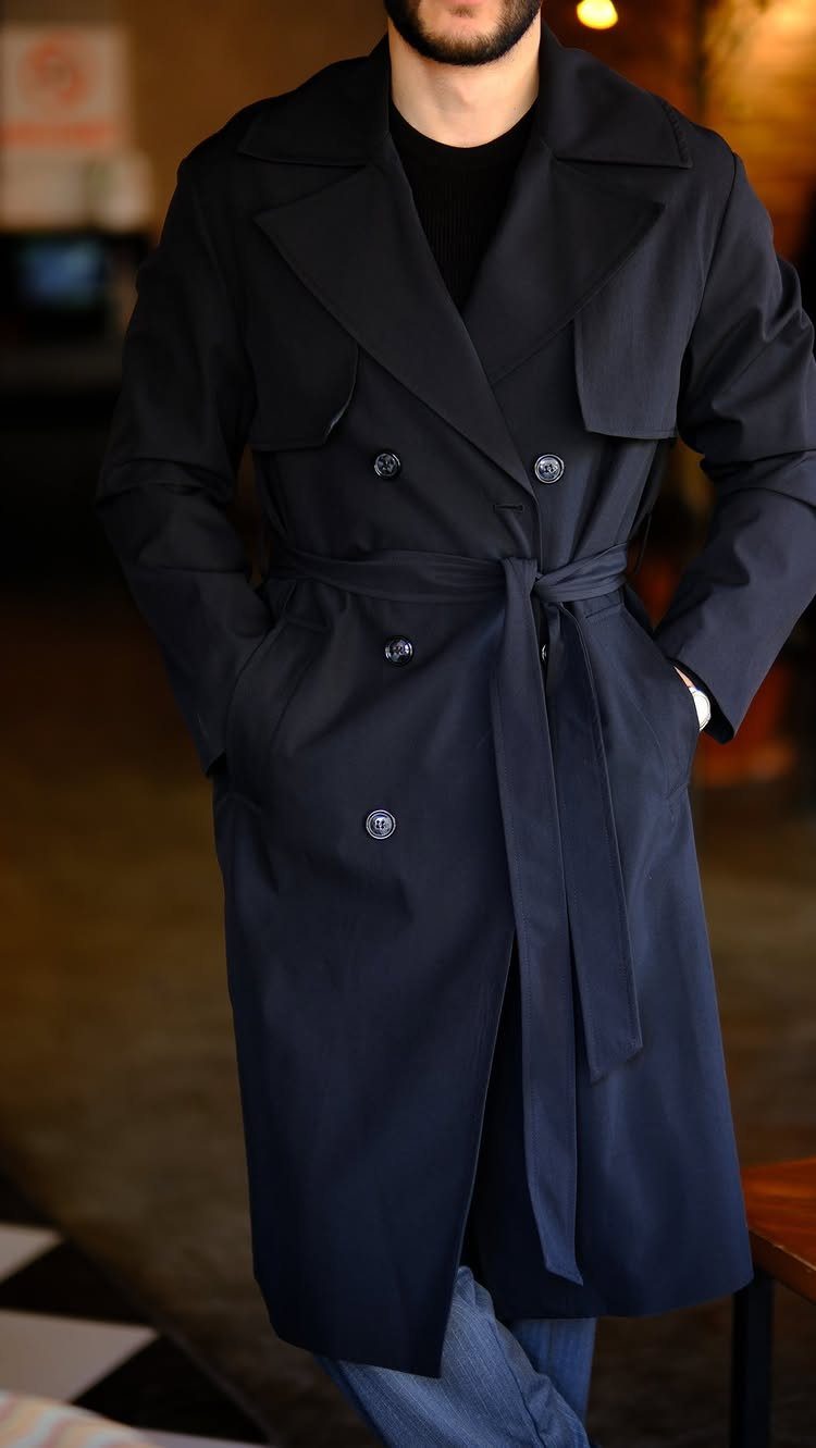 Man wearing a navy blue belted trench coat with blue jeans indoors