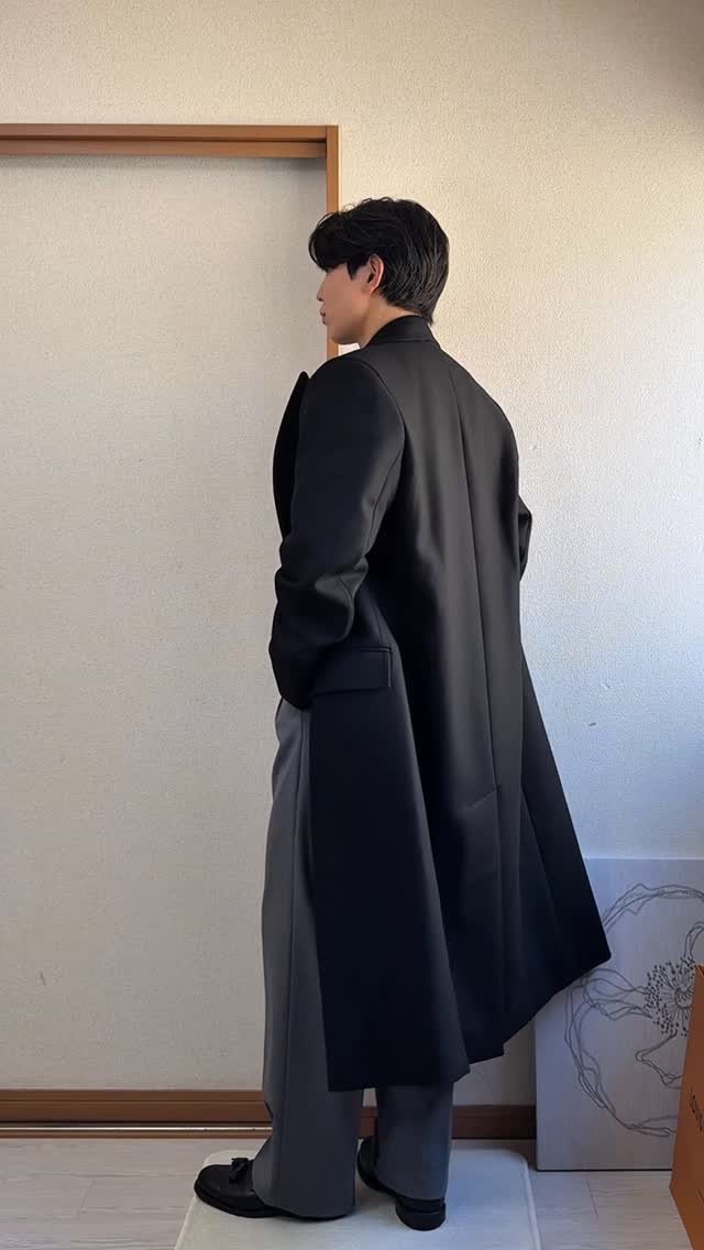 Man wearing a long black fall coat and grey trousers standing in profile.
