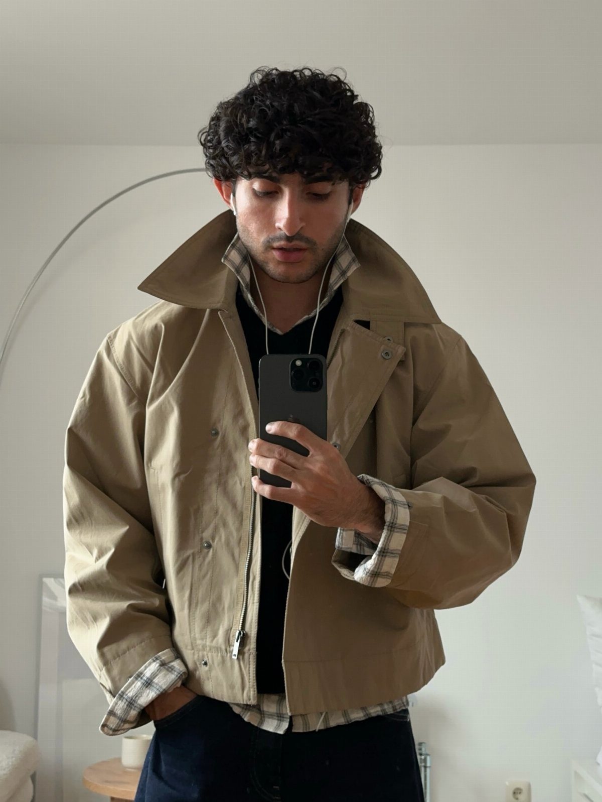 Man wearing a tan short jacket and plaid shirt taking a mirror selfie.