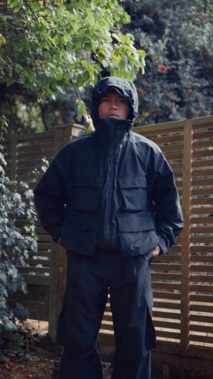 Person wearing a black hooded utility rain jacket and matching trousers outdoors.