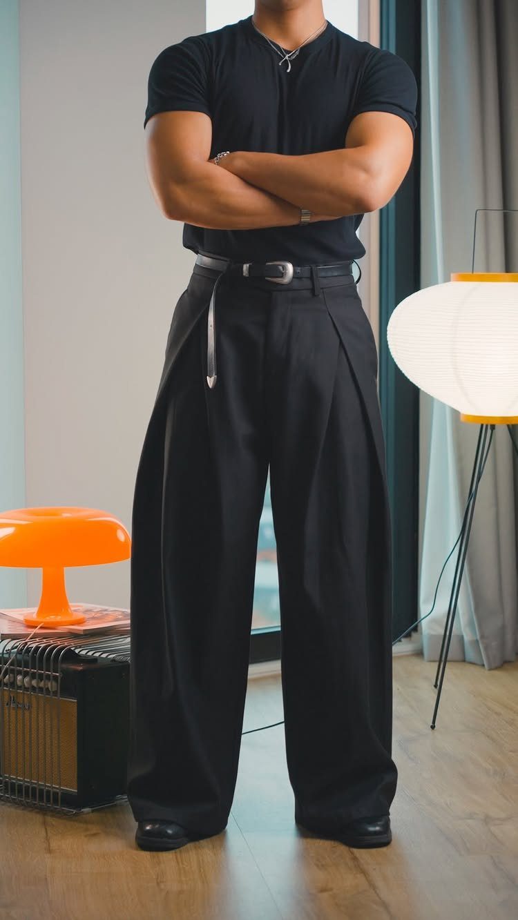 Man wearing a black short-sleeved shirt and wide-leg black trousers in a room.