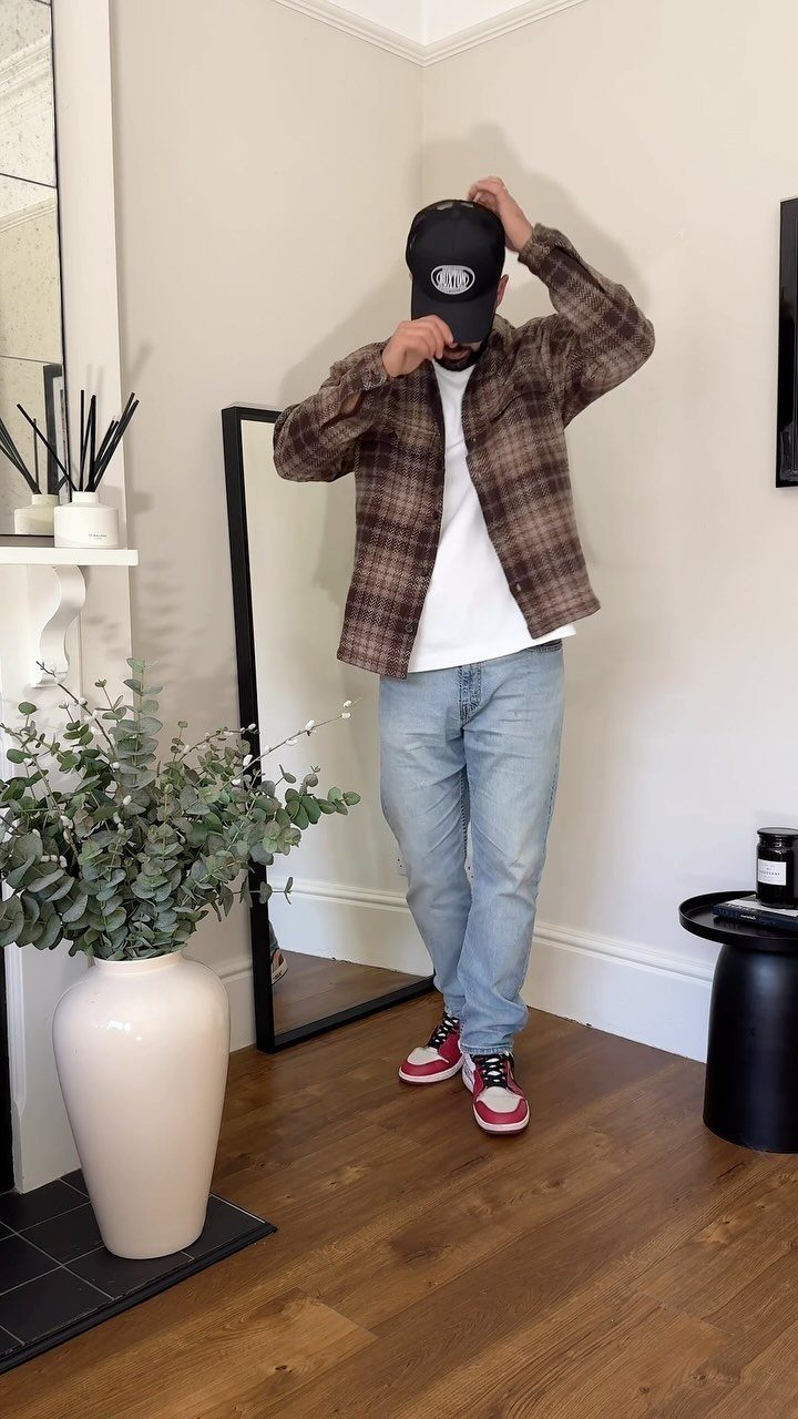 Man wearing a brown plaid shirt, white t-shirt, and light blue jeans indoors.