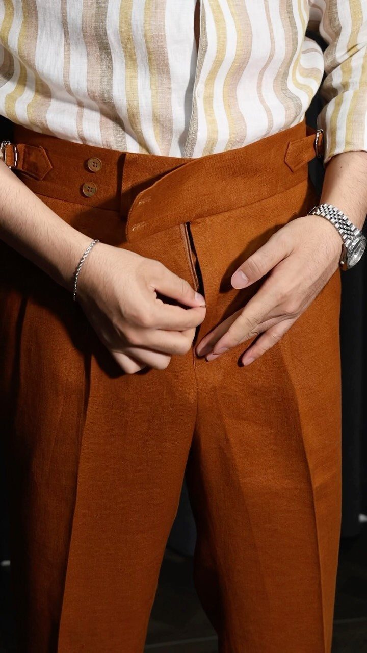 Rust-colored pleated trousers paired with a beige and white striped button-down shirt.