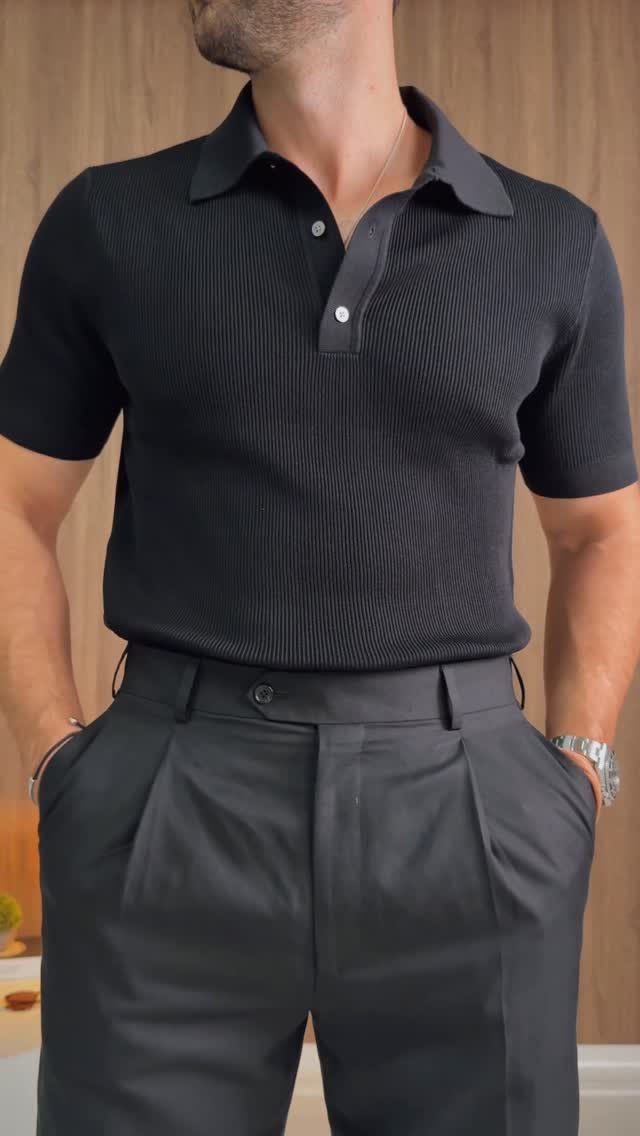 Man wearing a black knit polo shirt and black pleated trousers against wood wall