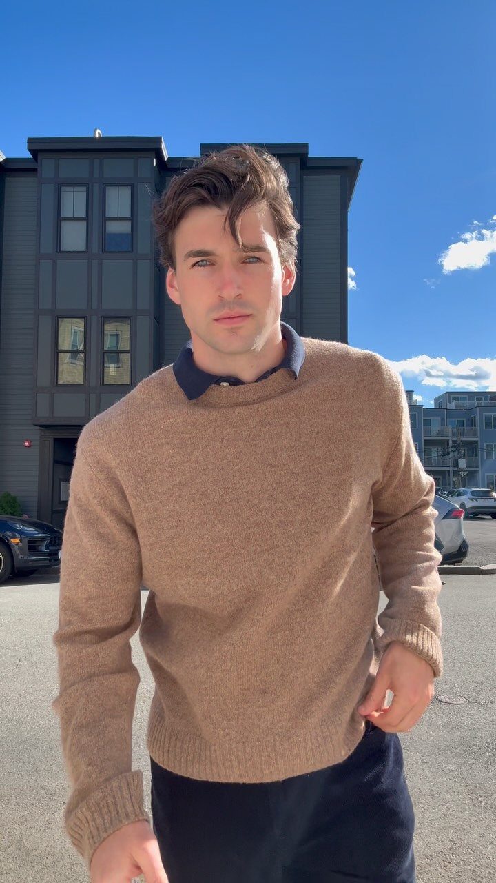 Man in a brown crewneck sweater and dark trousers standing before a grey building.