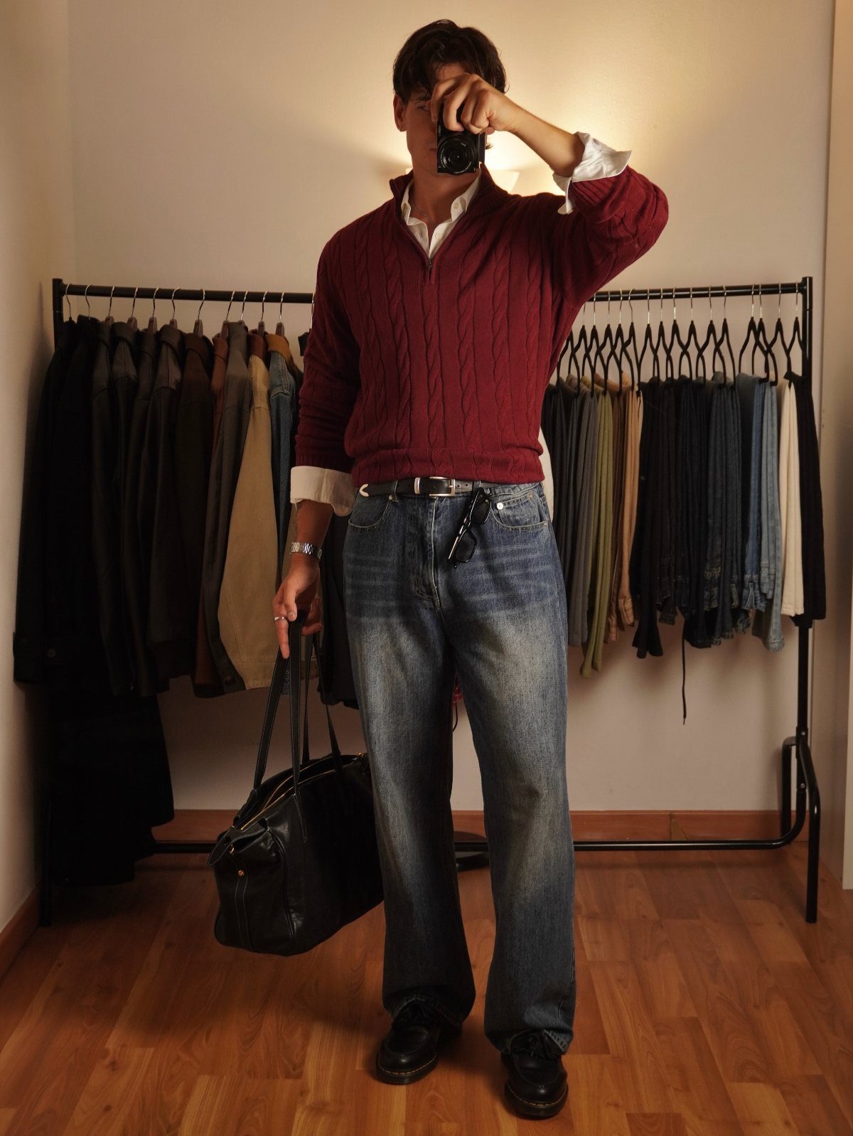 Man wearing a burgundy cable-knit sweater and blue jeans standing by a clothing rack.