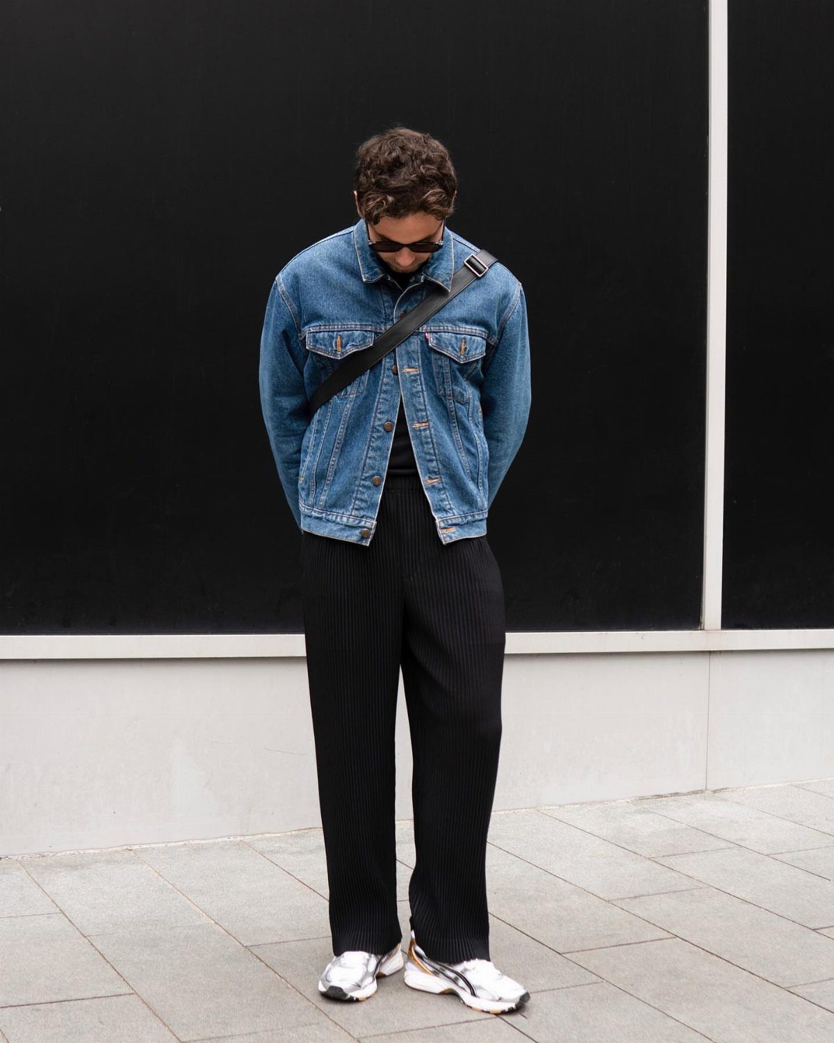 Person wearing a blue denim jacket and black wide-leg trousers against a black wall.