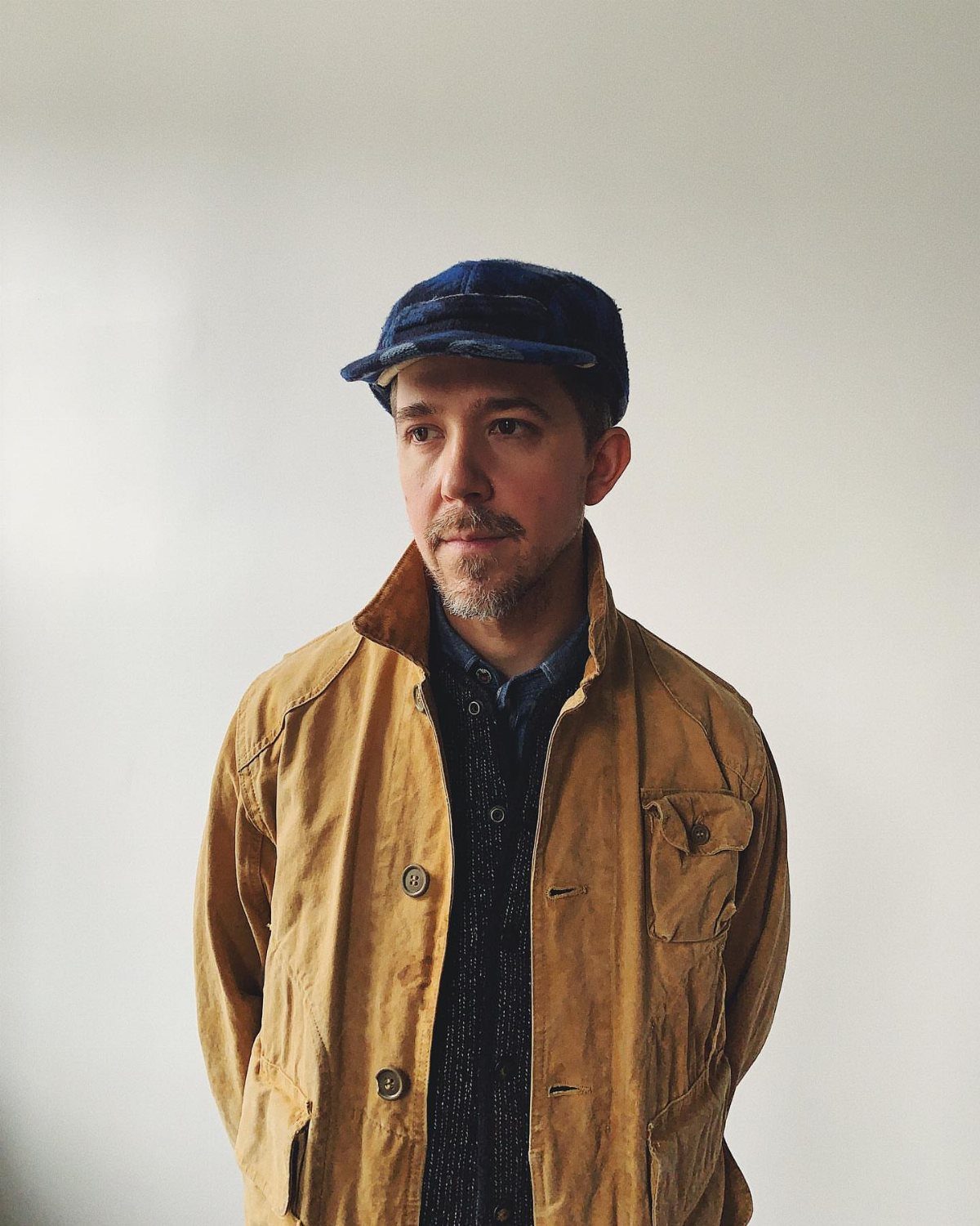 Man wearing a tan field jacket and blue flat cap against white background