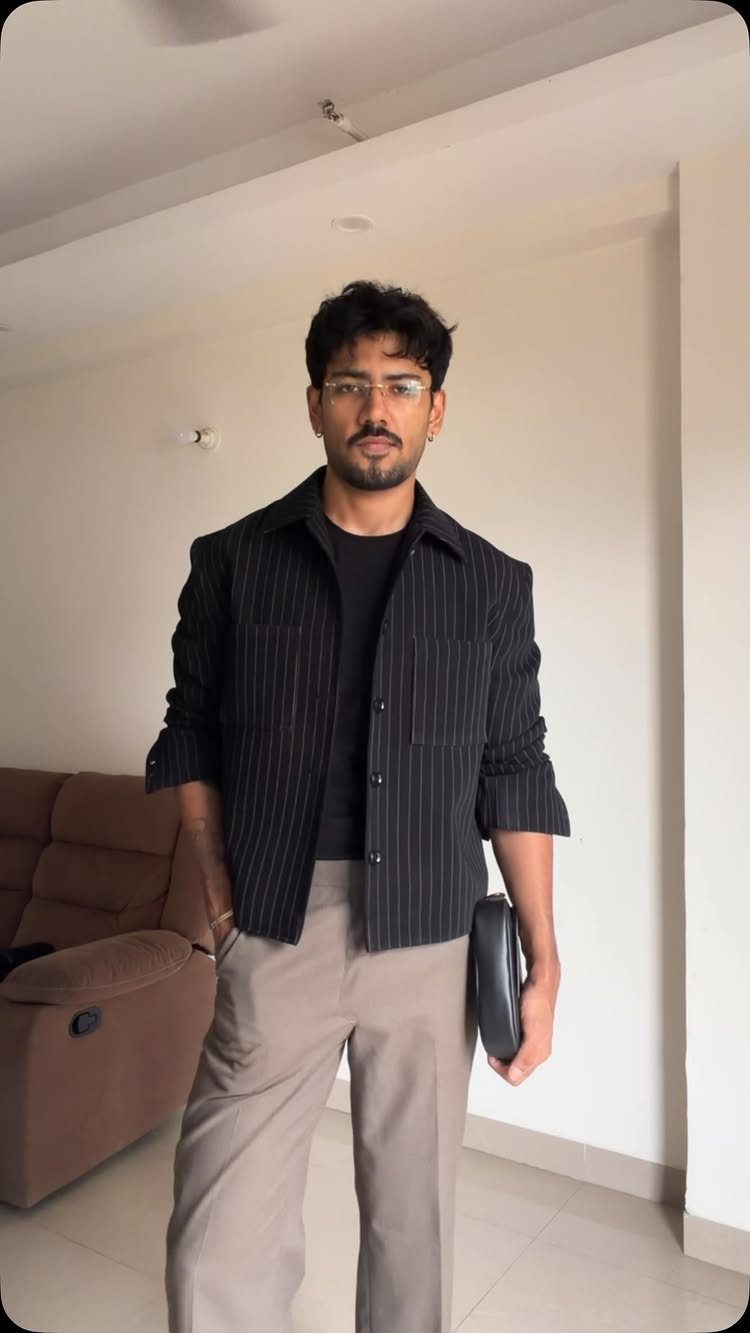 Man wearing a black pinstriped shirt and brown trousers holding a black clutch.
