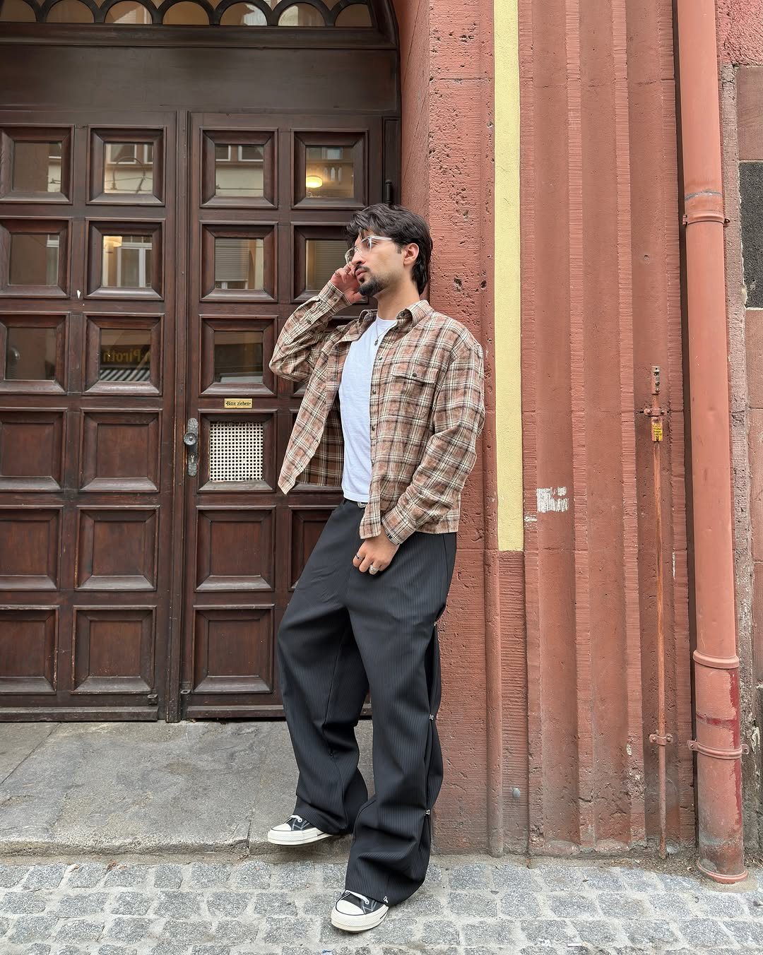 Man wearing a white t-shirt under a brown plaid flannel shirt and black trousers.