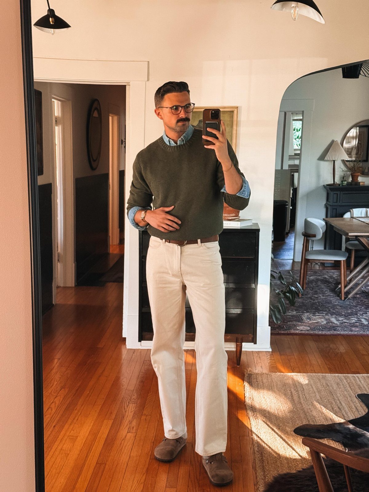 Man wearing an olive crewneck sweater over a blue shirt and white trousers.