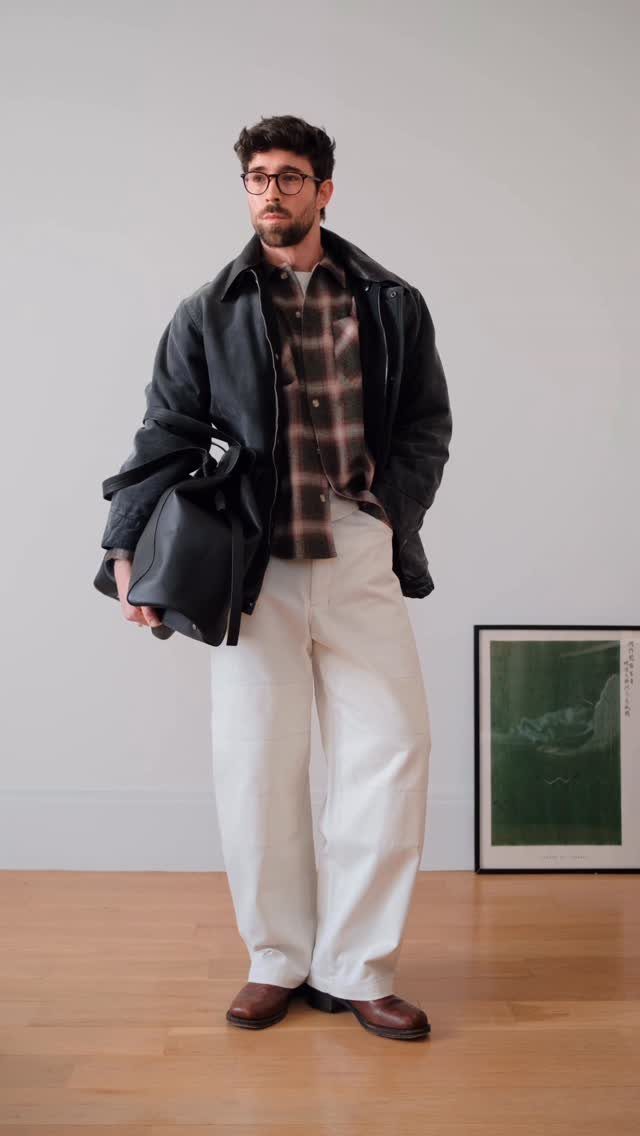 Man in black jacket, plaid shirt, and wide white trousers holding a black bag.