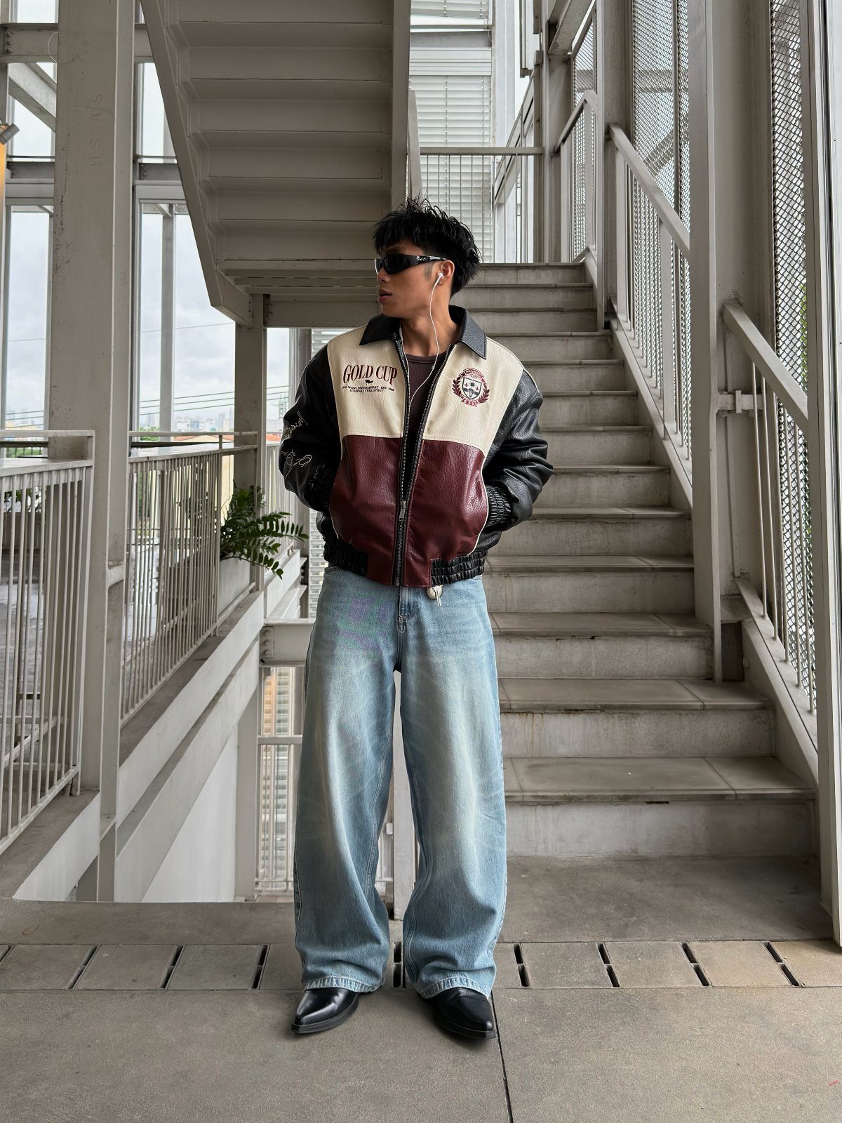 Person wearing a leather bomber jacket and wide-leg jeans standing by a staircase.