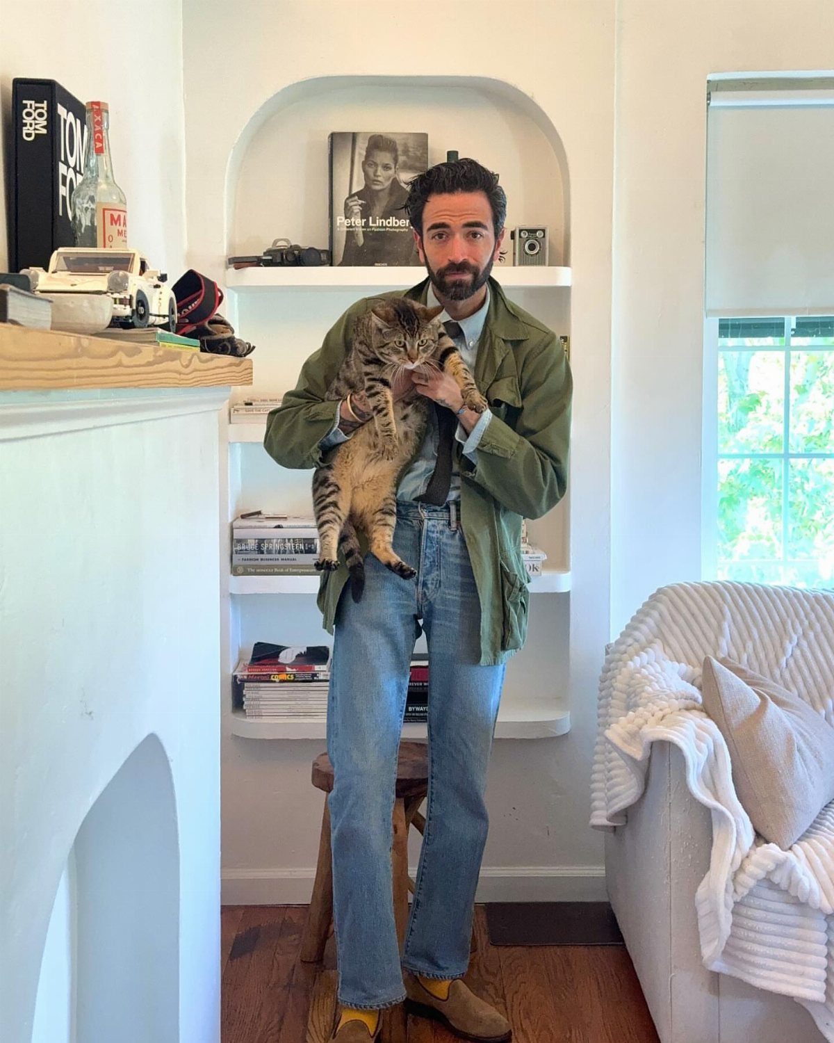 Man in olive green jacket and blue jeans holding a tabby cat indoors