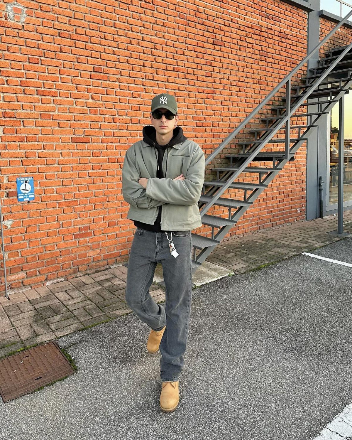 Man wearing a sage green jacket and gray jeans standing by a brick wall