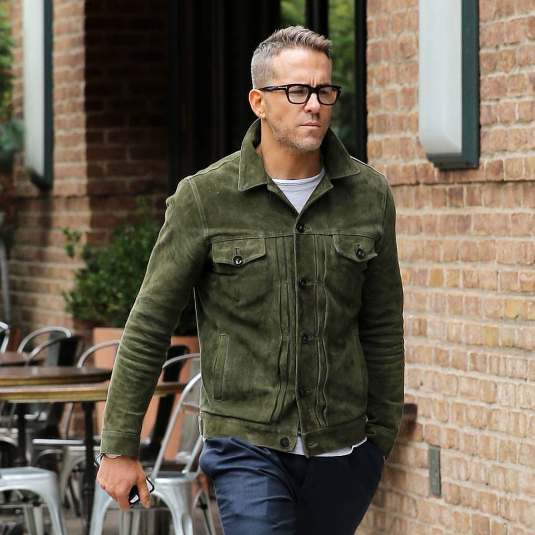 Man wearing an olive green suede jacket, white shirt, and navy trousers outdoors.