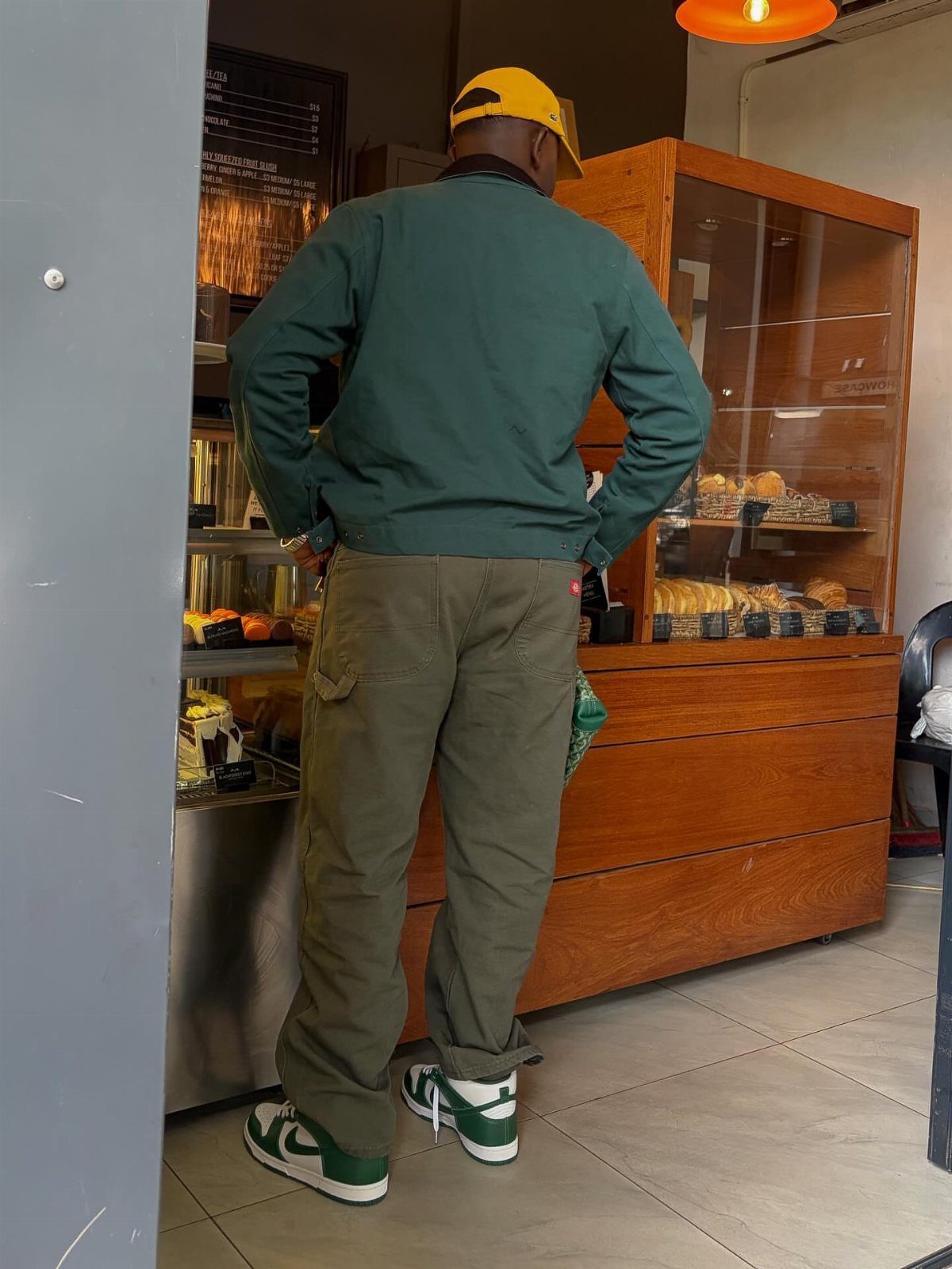 Person from behind wearing a green jacket, olive cargo pants, and yellow cap.