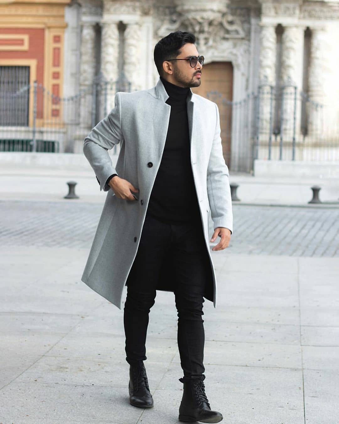 Man in light grey overcoat, black turtleneck, and black skinny jeans standing outdoors.