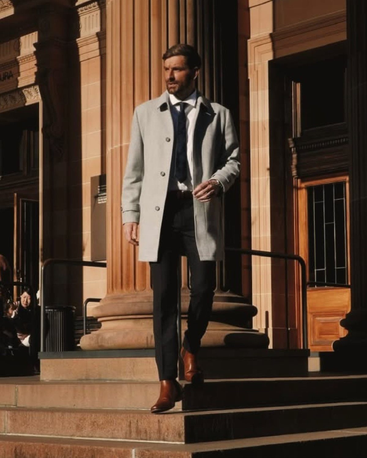 Man in a grey overcoat and black trousers wearing brown leather boots.