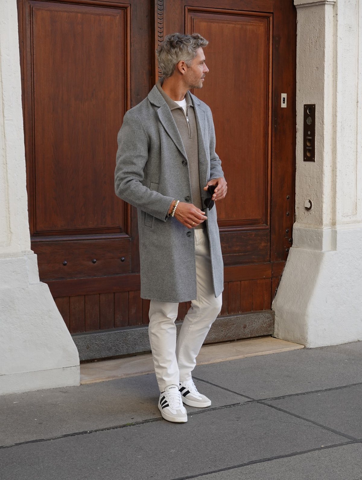 Man wearing a grey overcoat, white trousers, and white low top sneakers.