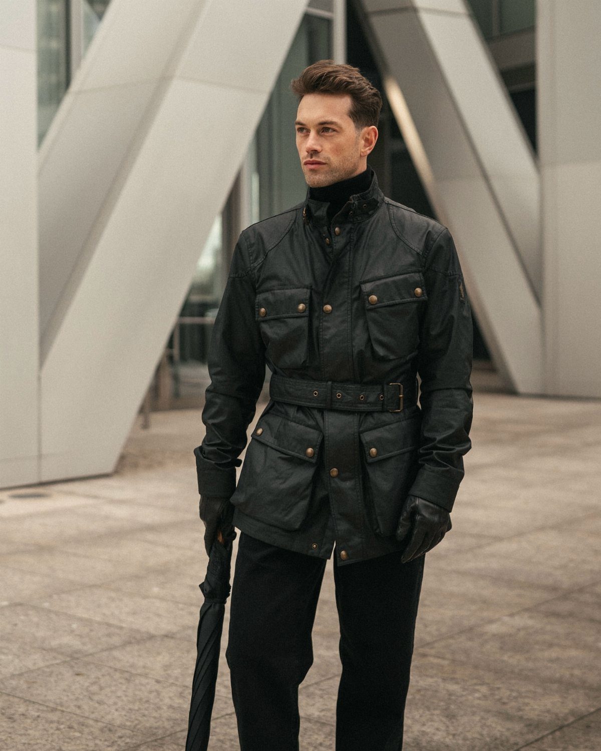 Man in a tailored black belted coat and gloves holding a black umbrella.