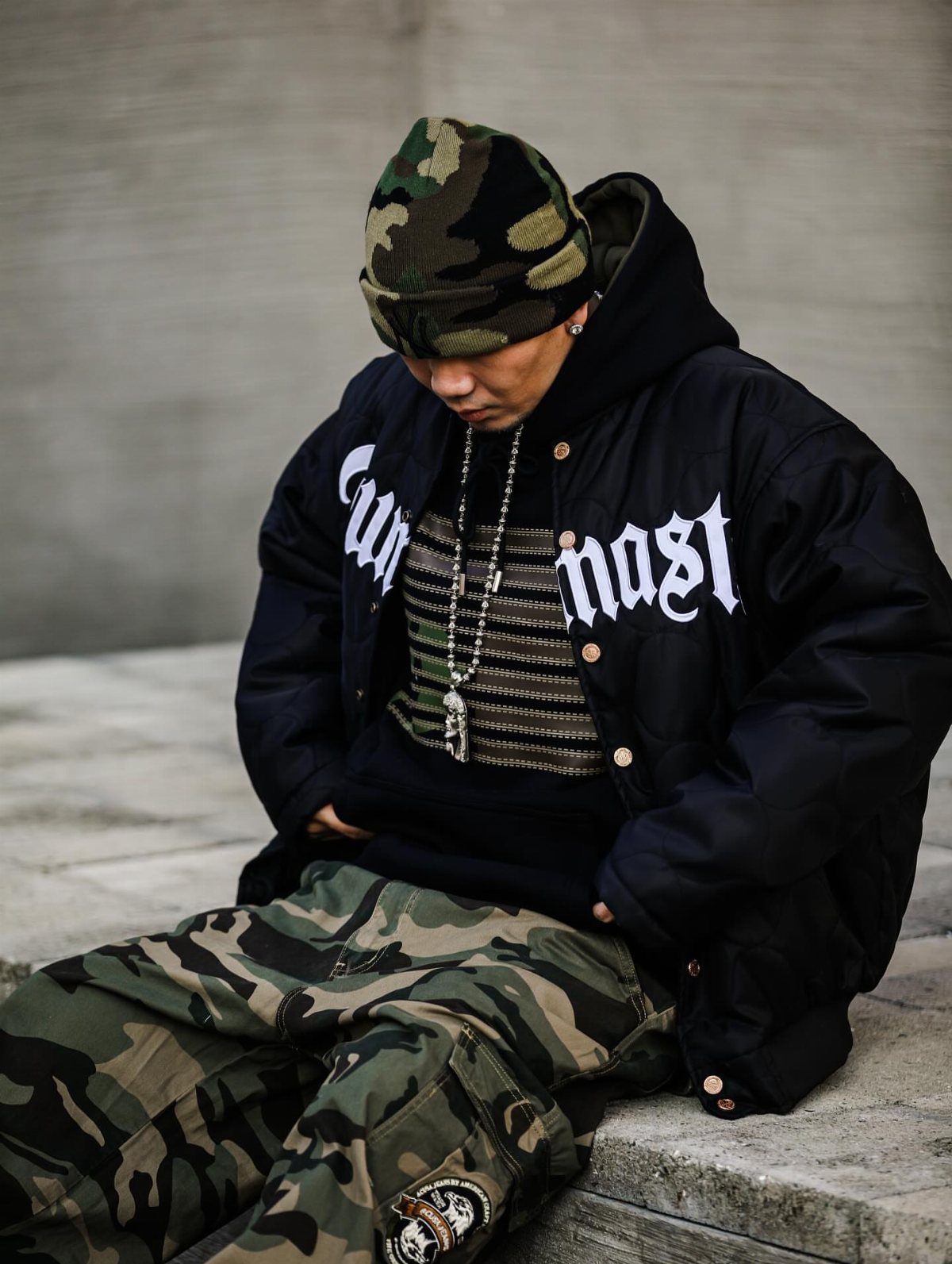 Person in oversized black gothic text jacket and camouflage clothing sitting on concrete steps