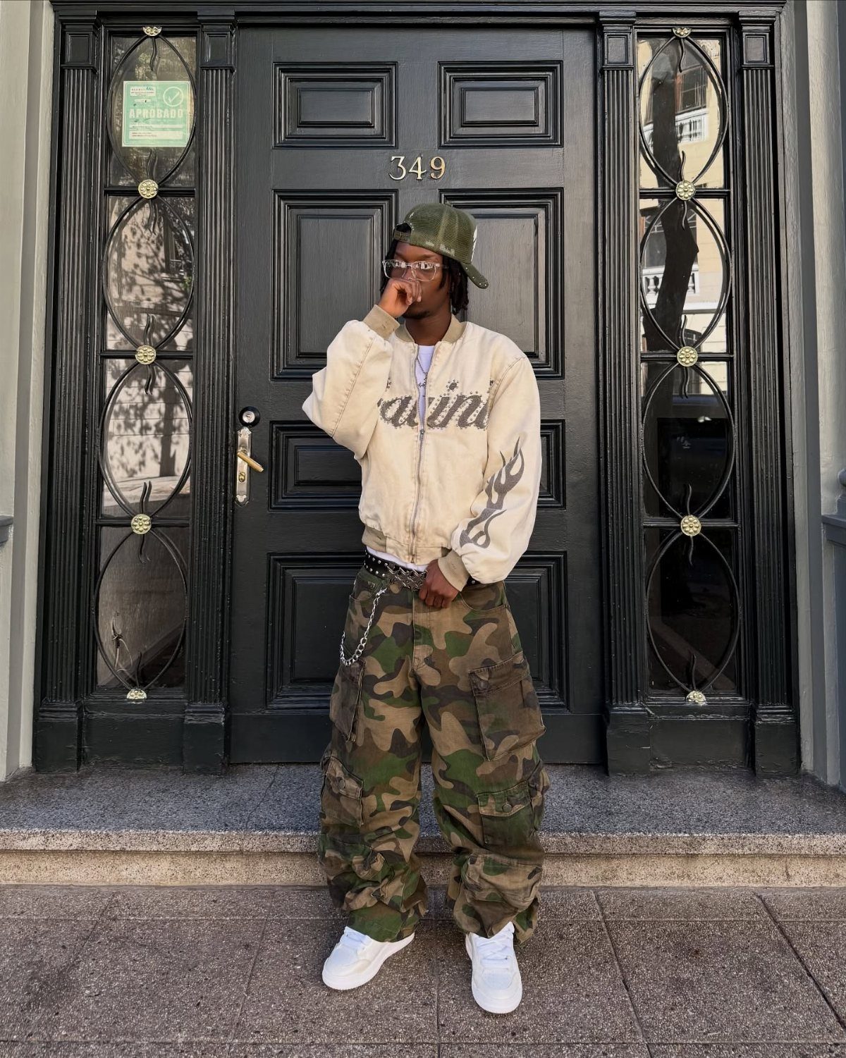 Person in beige bomber jacket and camouflage cargo pants standing before black door