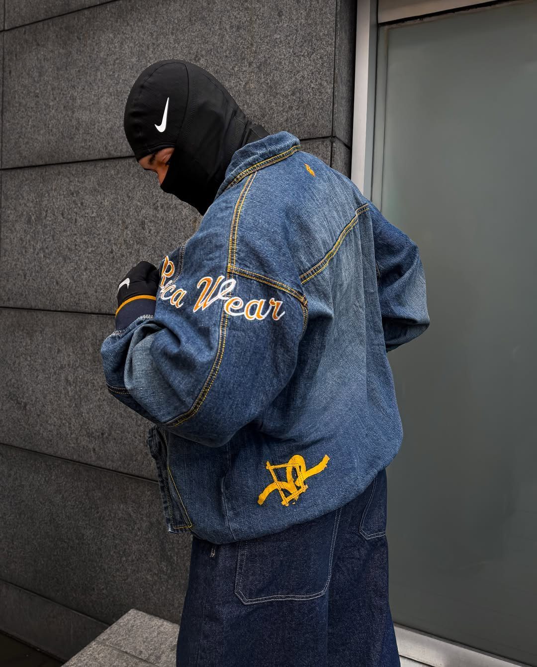Person in a black balaclava wearing an oversized blue denim jacket with yellow embroidery.