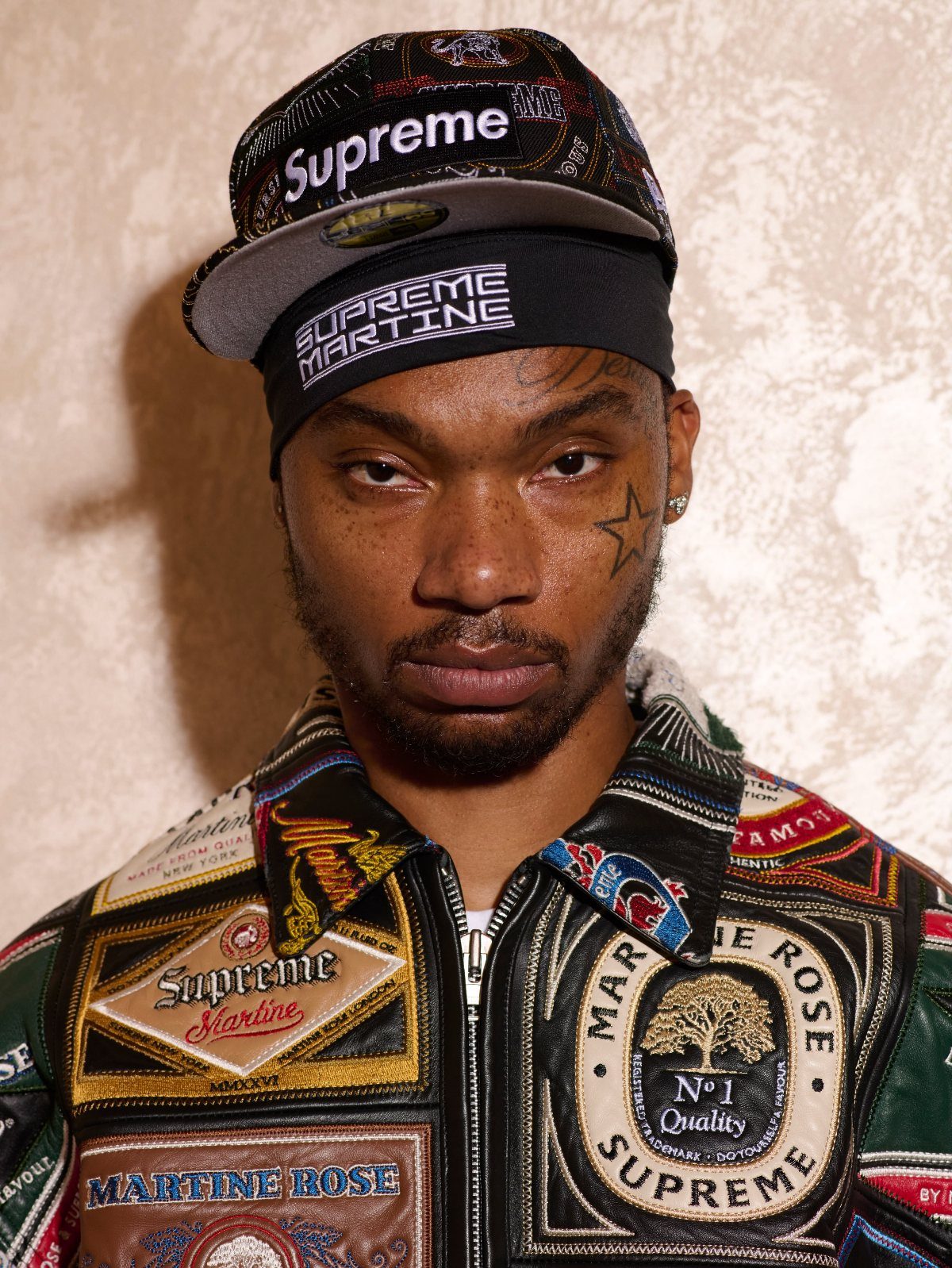 Man wearing a Supreme cap and a leather jacket with multiple colorful patches.