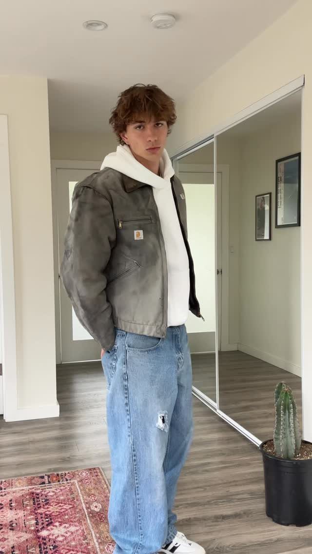 Young man wearing an olive green work jacket, white hoodie, and baggy ripped jeans.