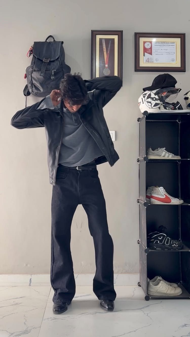 Man in black leather jacket and flared trousers standing in a room.