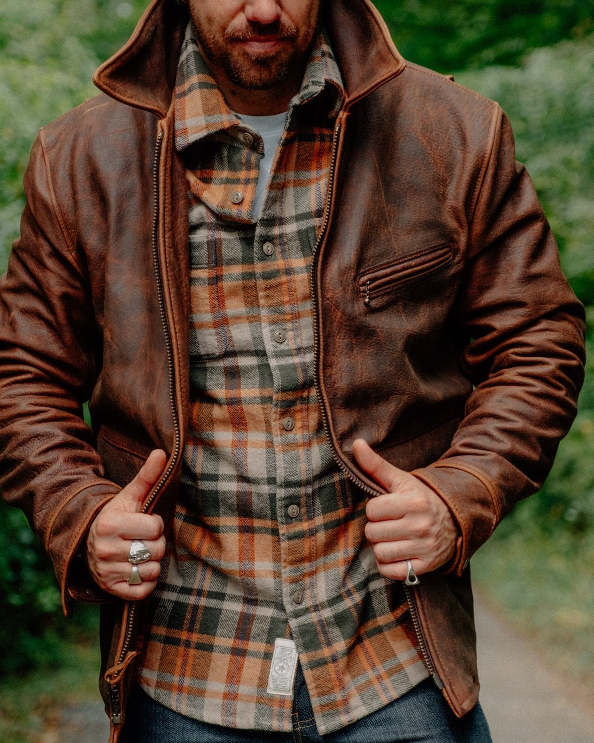Man wearing a brown leather jacket over an orange plaid flannel shirt outdoors.