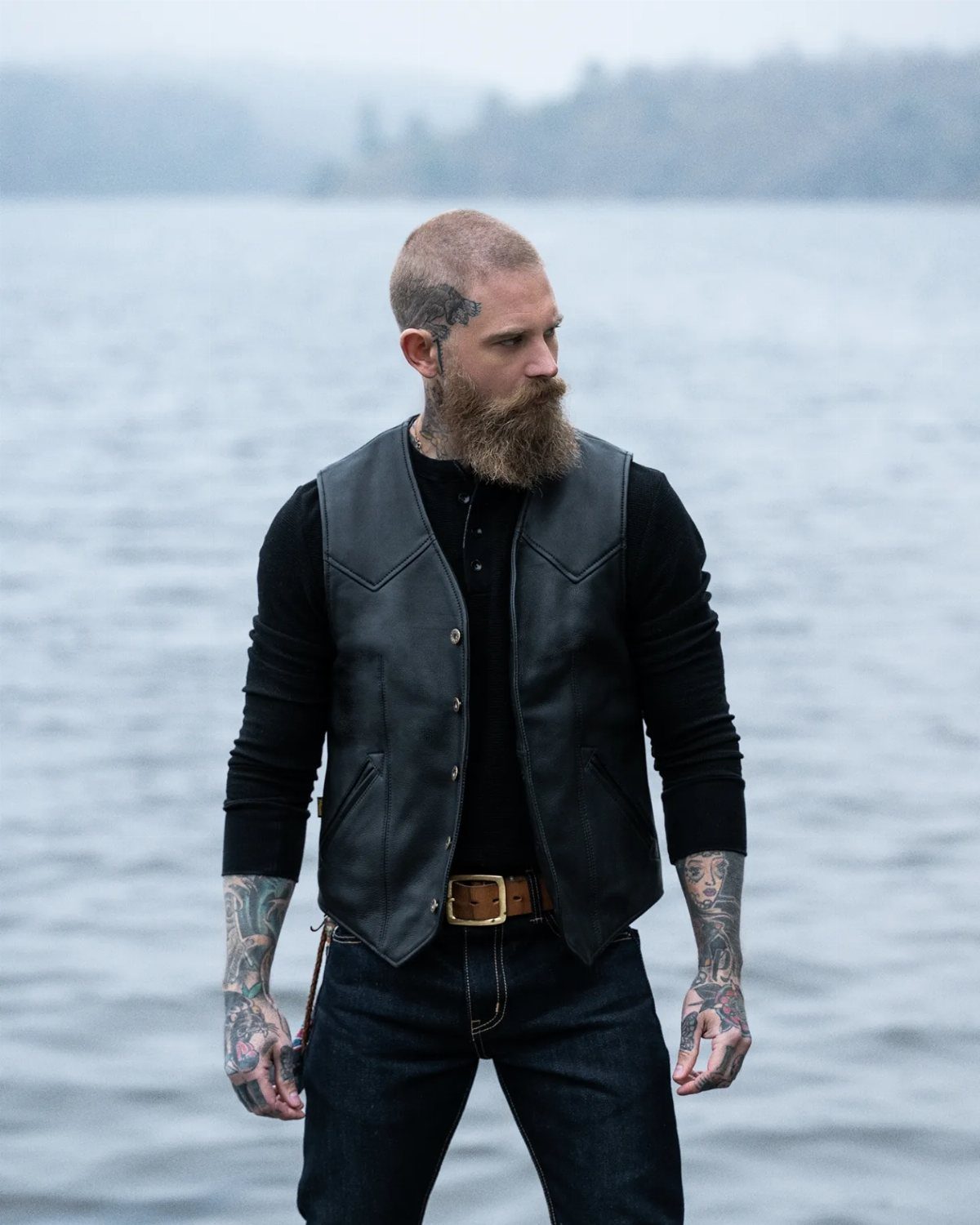 Bearded man wearing a black leather vest over a black sweater and jeans.
