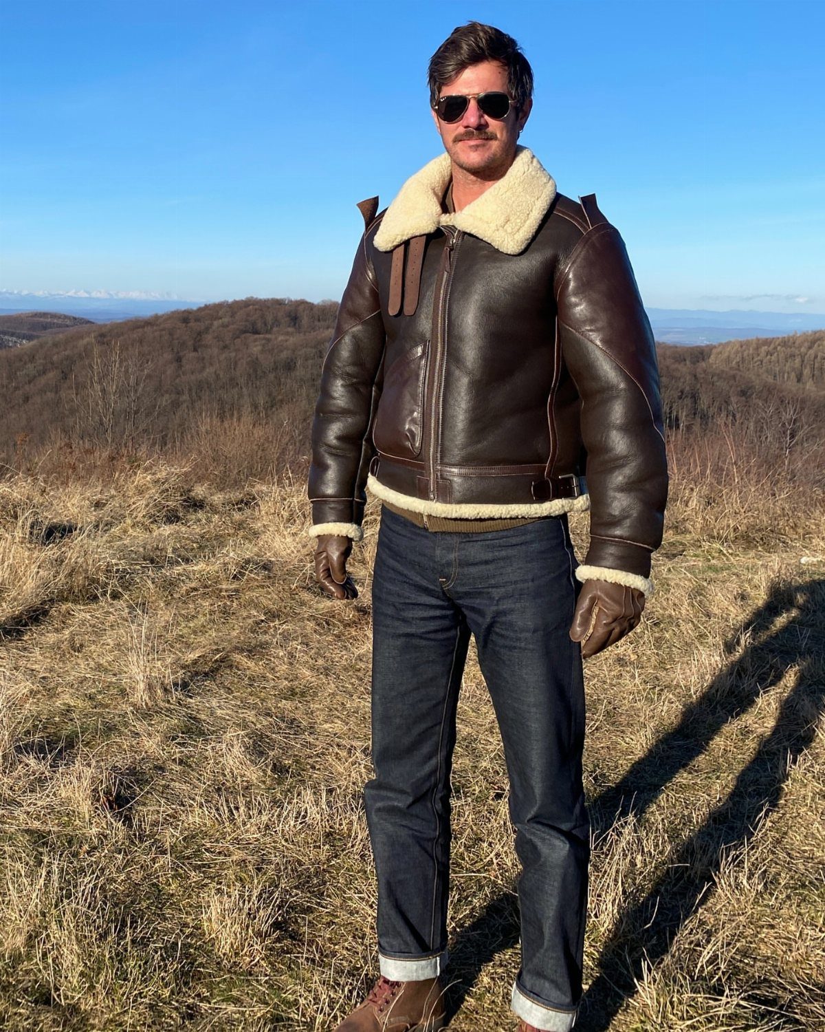 Man wearing a brown leather shearling flight jacket and dark blue jeans outdoors.