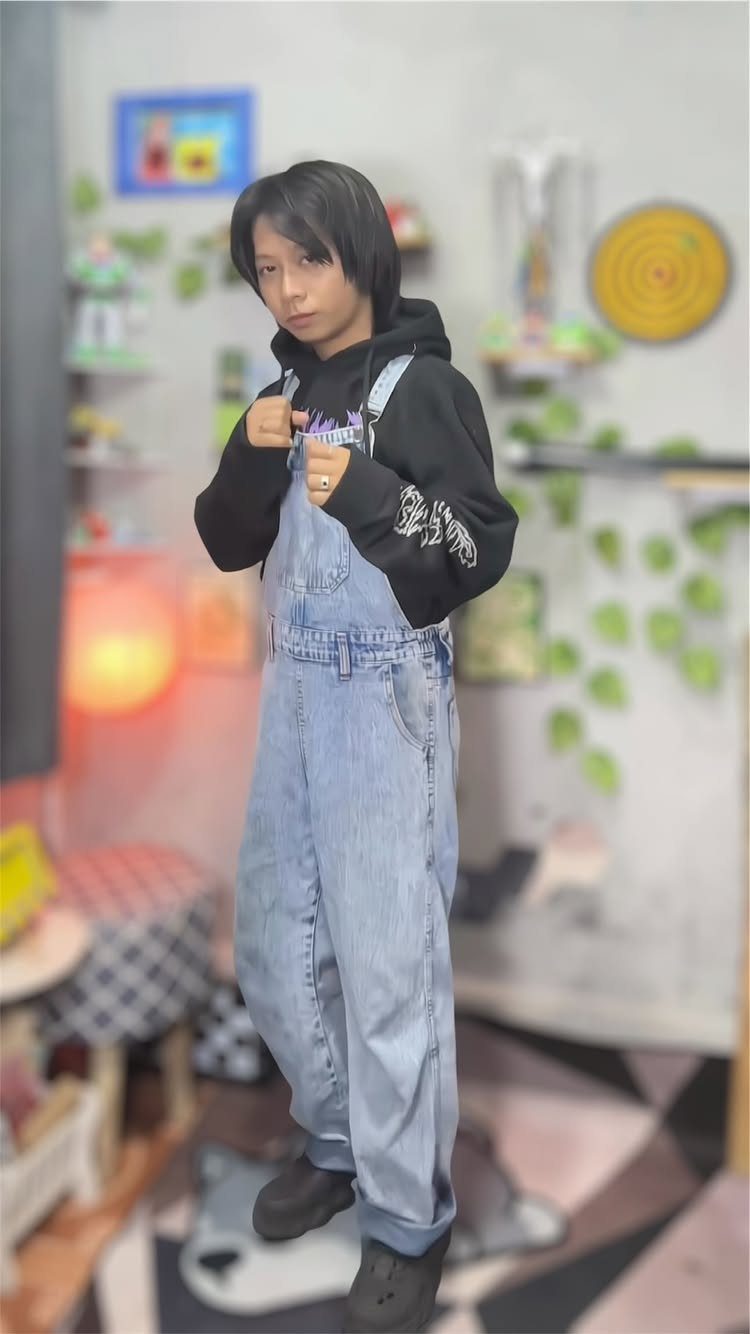 Person wearing light blue denim overalls over a black hoodie in a room.