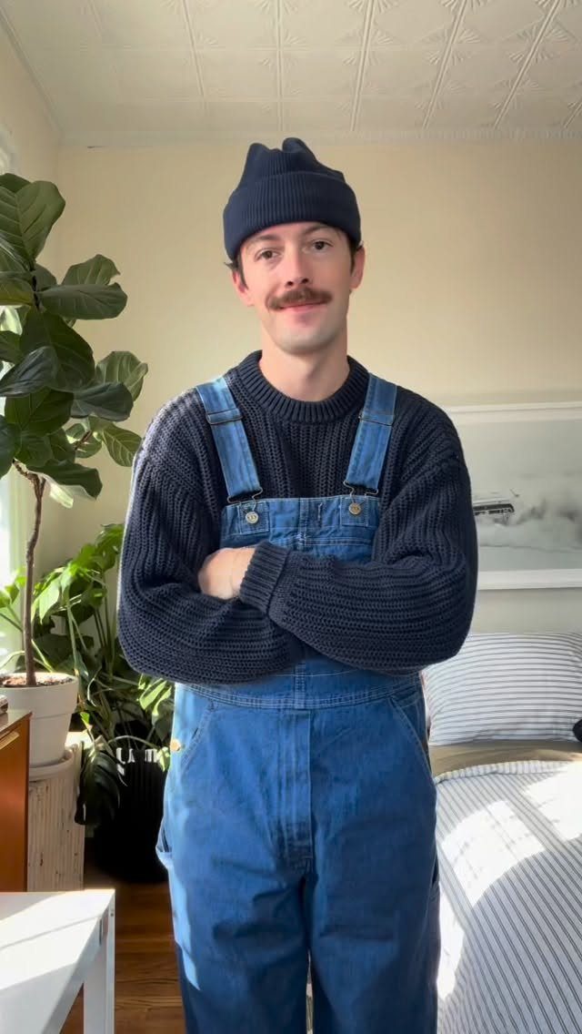 Man wearing blue denim overalls, a navy ribbed sweater, and a navy beanie.