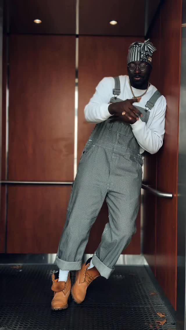 Man in striped overalls and white shirt leaning against a brown wooden wall.