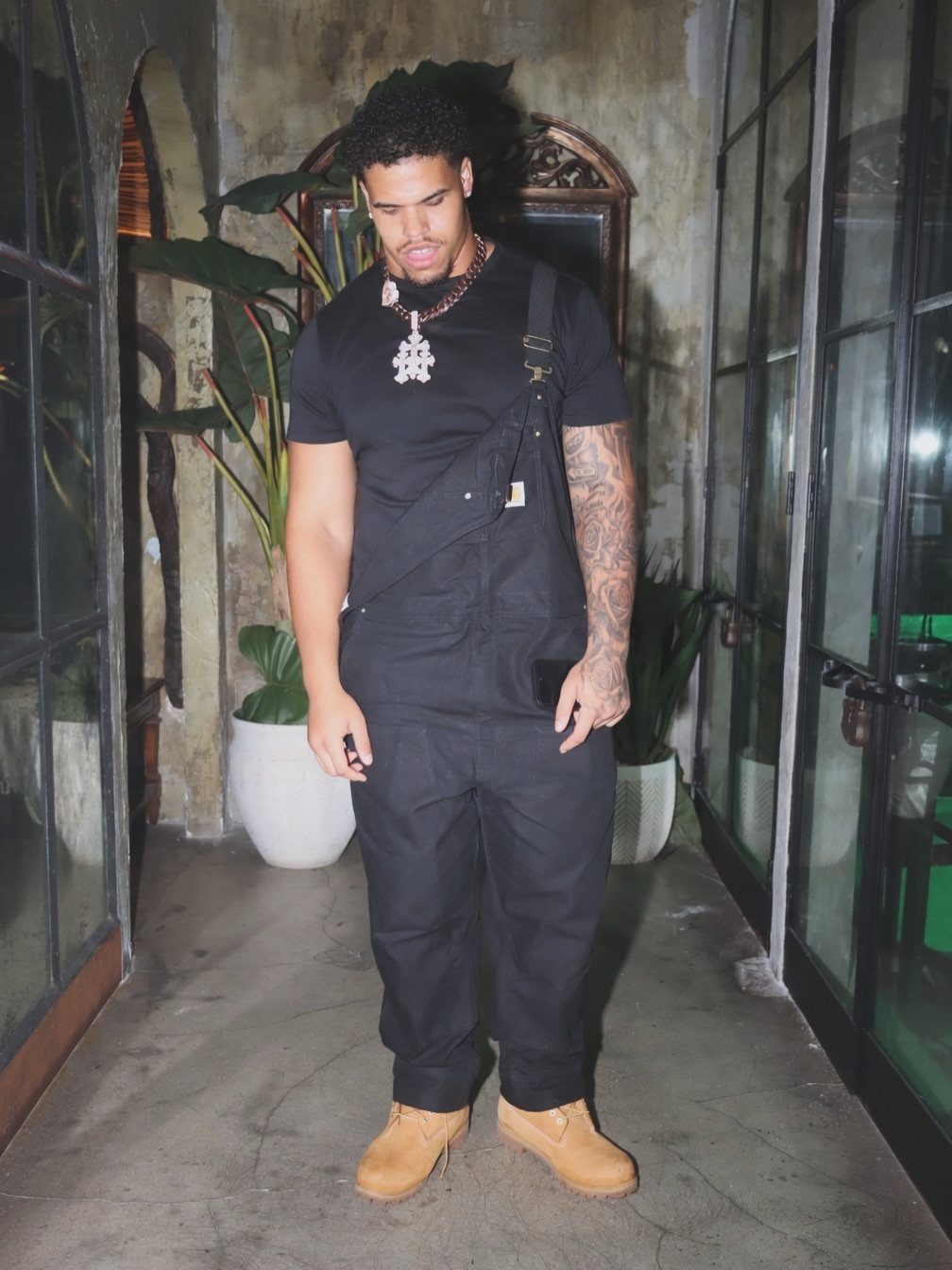 Man wearing black overalls, a black t-shirt, and tan work boots in a hallway.
