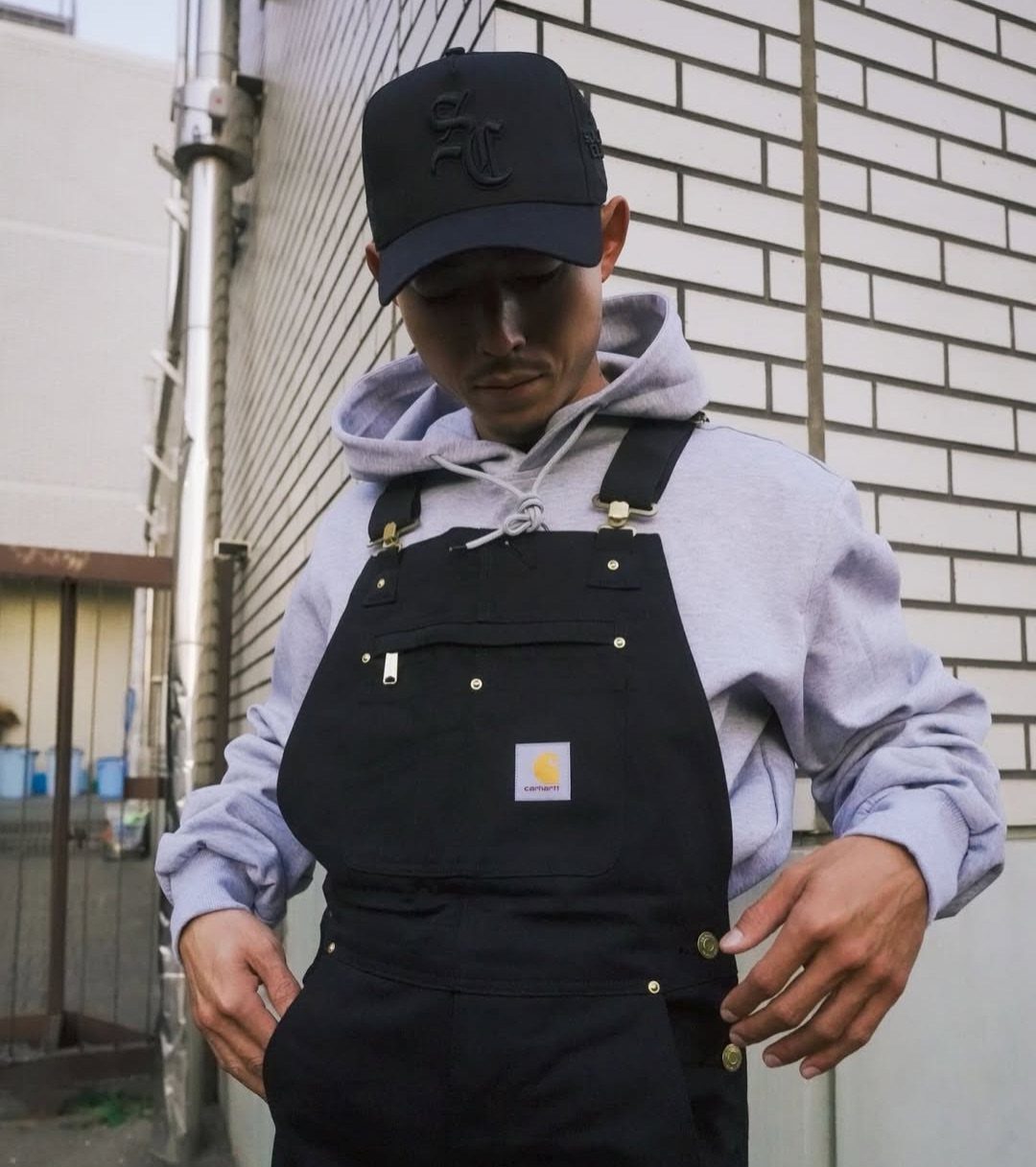 Man wearing black overalls over a grey hoodie and a black baseball cap.