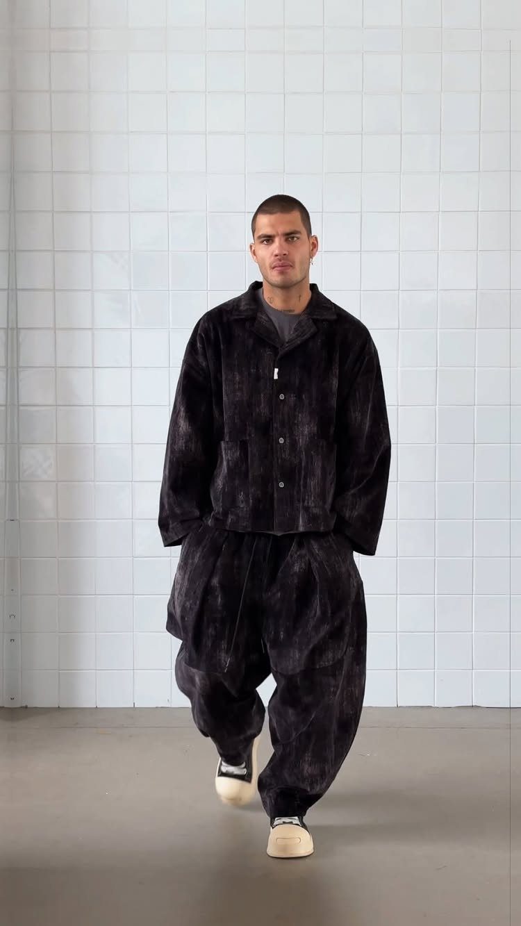 Man wearing an oversized dark corduroy jacket and matching trousers against white tiles.