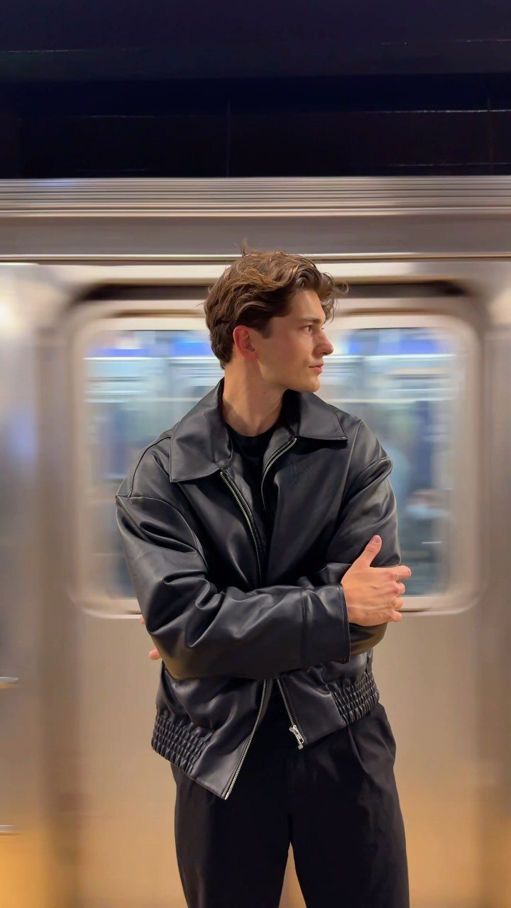 Young man wearing a black leather jacket and trousers standing by a subway train.