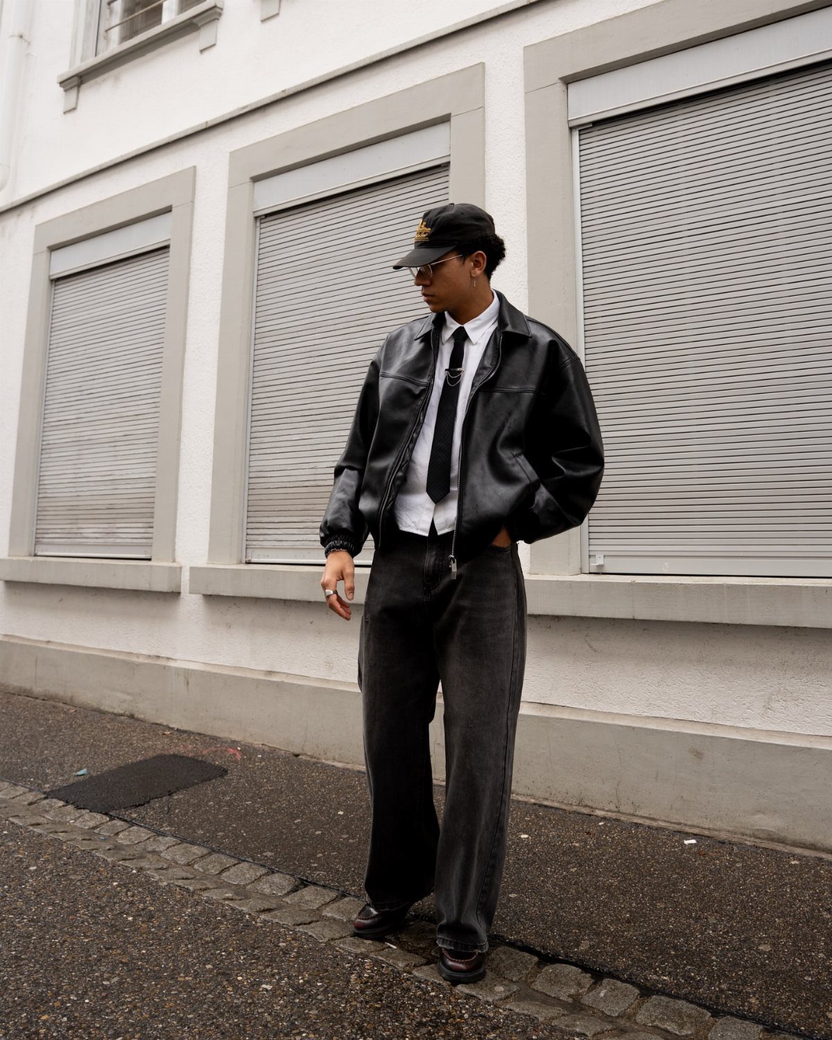 Man wearing a black leather jacket, white shirt, black tie, and baseball cap.