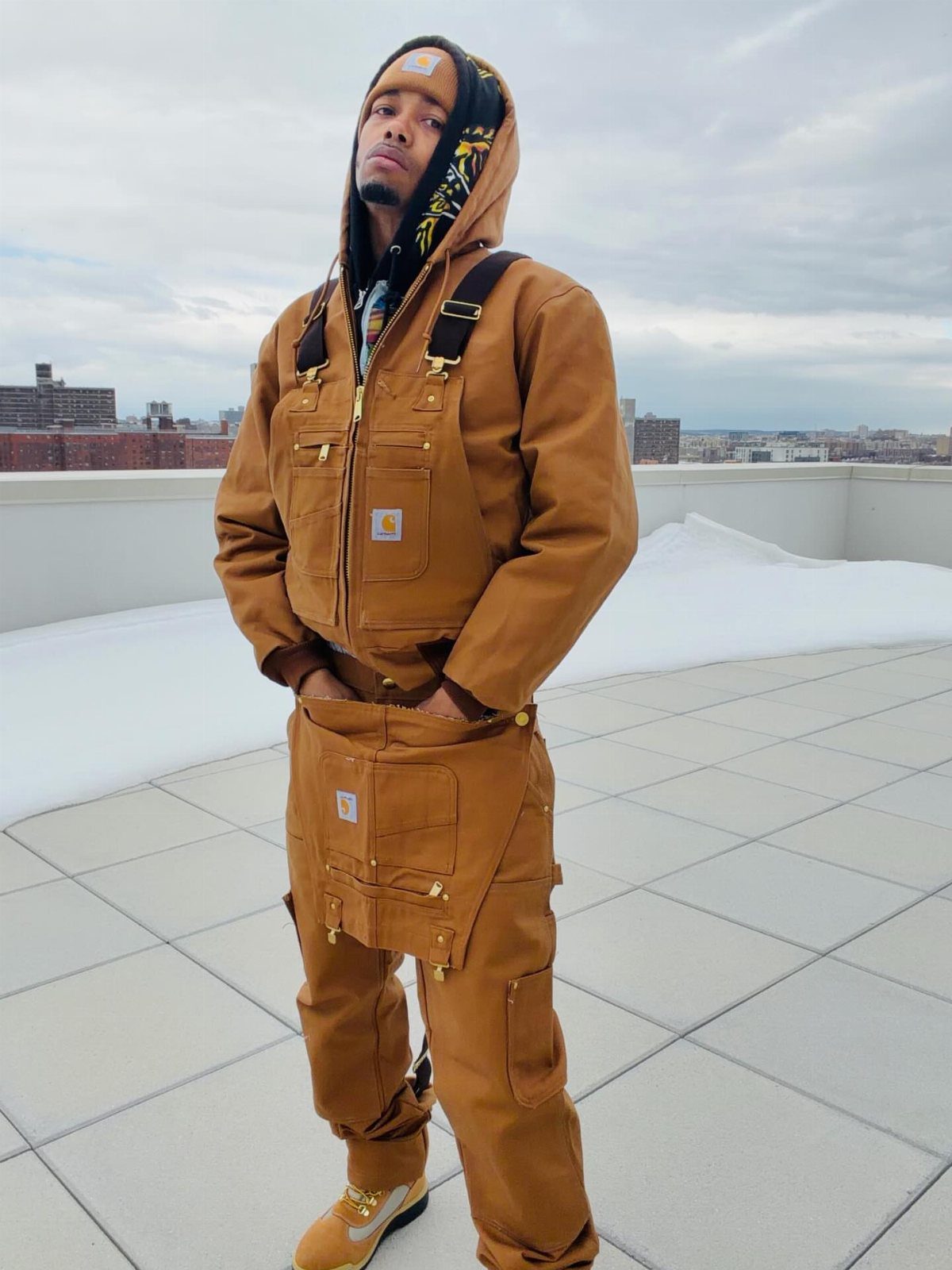 Man wearing brown dungarees, a matching work jacket, and a black hoodie on rooftop.