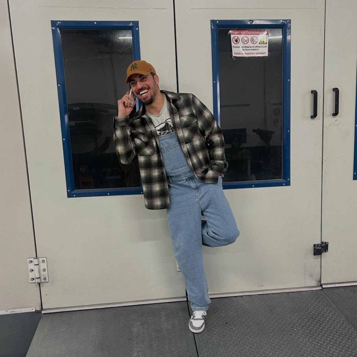 Man wearing light blue denim dungarees and a plaid flannel shirt leaning against wall.