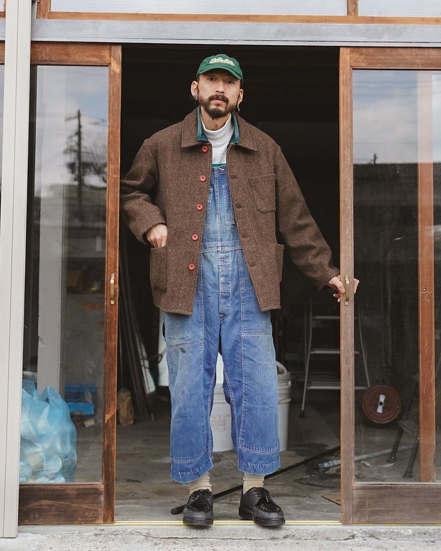 Man wearing blue denim dungarees with a brown wool jacket and green cap.