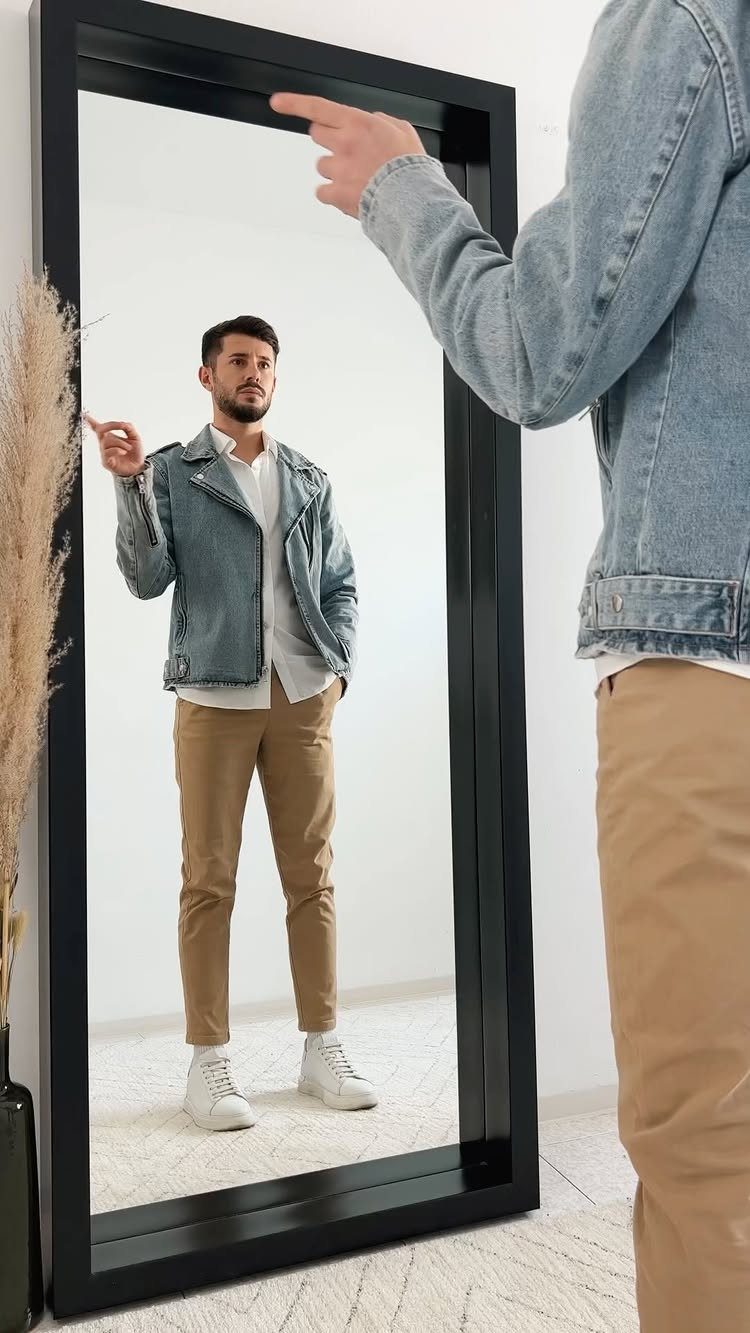 Man wearing a light denim jacket and tan chinos reflected in a mirror.