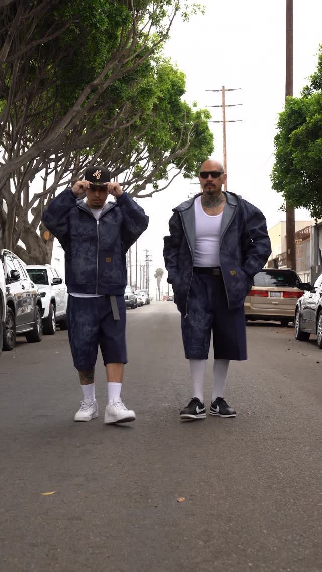 Two men wearing matching dark blue denim jackets and denim shorts on a street.