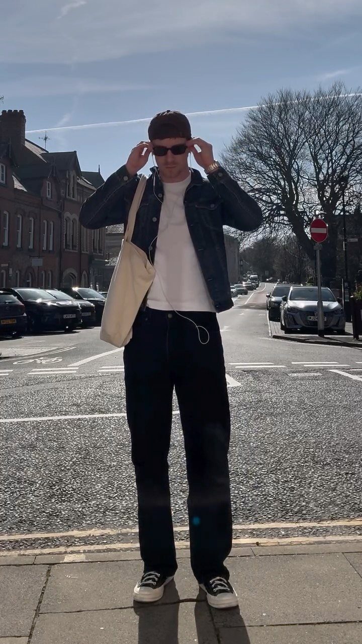 Man wearing a dark denim jacket, white t-shirt, and black trousers on a street.